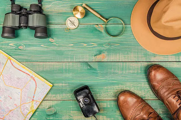 Tourism concept. Magnifying glass, compass, city map, binoculars, brown shoes, fedora hat and old film camera on green wooden background