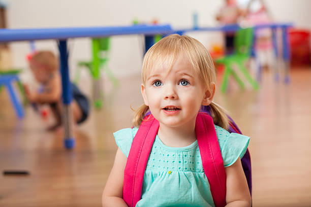 Beautiful Caucasian toddler girl enjoys her preschool class. She is wearing a pink backpack. Her blond hair is in pigtails.