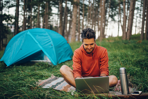 Vintage toned image of a young man camping in the forest, in front of the tent. He is looking at his laptop with a SIM card internet, checking e-mail while far away from the city and urban lifestyle. Taken in the Dinaric Alps mountain range in the Southeastern Europe