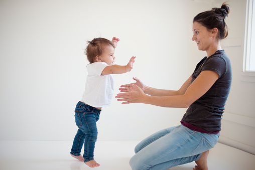 A mother and her baby girl. The baby girl is walking her firs steps towards her mother. The happy mother is tending her arms to her child. They are smiling to each other. They are having fun. The baby girl is wearing a white shirt and blue jeans. The mother is wearing a grey shirt and light jeans. The background is white. Horizontal indoors shot with copy space. This was taken in Quebec, Canada.