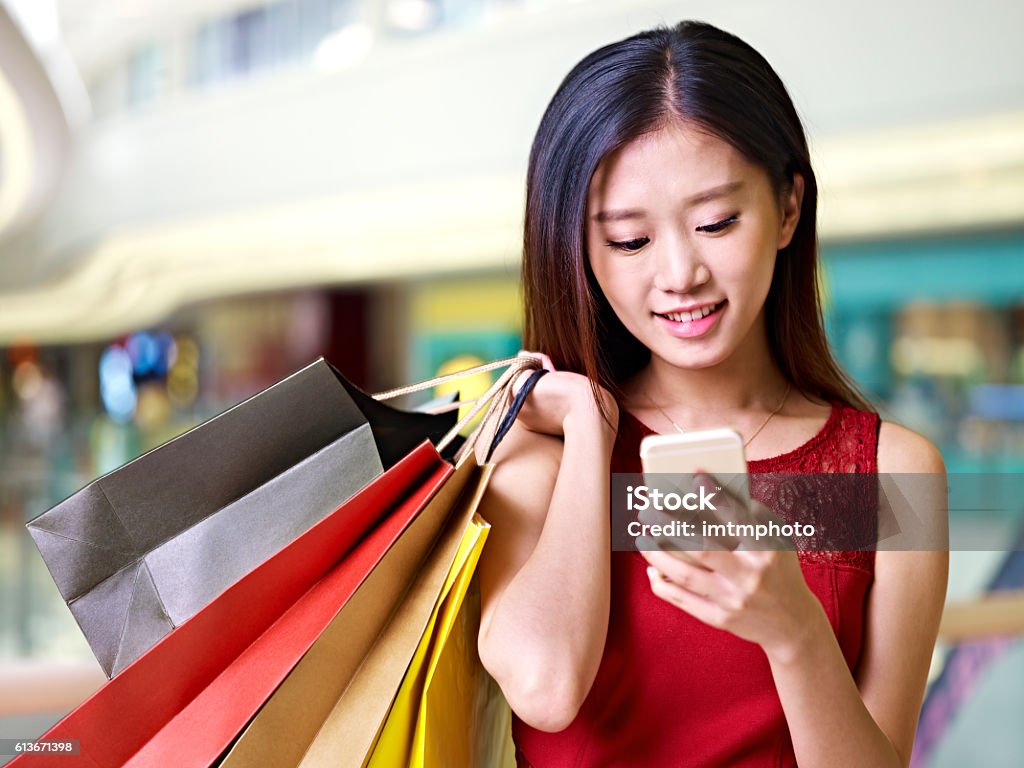 young asian woman using cellphone during shopping beautiful young asian woman carrying colorful paper bags on shoulder looking at mobile phone in modern shopping mall China - East Asia Stock Photo