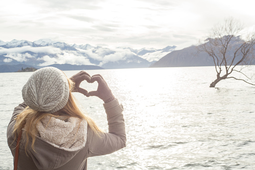 Young woman makes a heart shape finger frame on lake mountain landscape. 