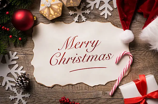 High angle view of Santa Claus letter with Merry Christmas text on it. Top view of merry Christmas xmas wish list with little gift present and santa claus hat on wooden background.