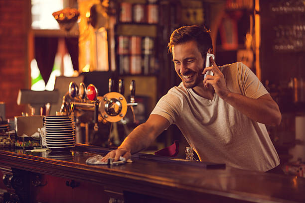 Happy bartender cleaning bar counter and talking over cell phone. Young smiling barista making a phone call while wiping a bar counter. bar cleaning stock pictures, royalty-free photos & images