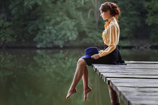 Side view of dreaming girl sitting on jetty forest and river on background. Beautiful woman sitting by the lake. Sad girl depressed sitting on bridge. Lonely woman sitting on a wooden bridge