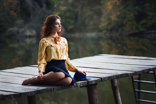 Dreaming girl with curly hair sitting on wooden bridge forest and river on background. Beautiful woman sitting by the lake. Sad girl sitting on bridge. Lonely woman sitting on a wooden bridge. Portrait of young woman on autumn background