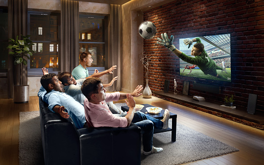 :biggrin:A group of young male friends are shocked while watching Soccer game on TV. They are sitting on a sofa in the modern living room. The TV set is on the loft brick wall. It is evening outside the window.
