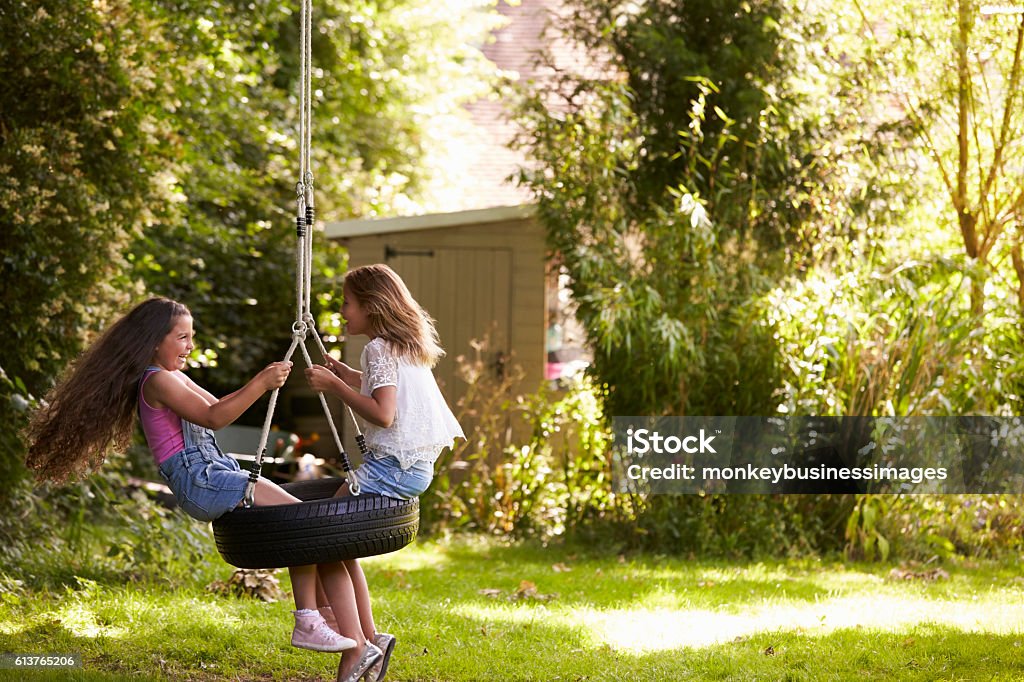 Two Girls Playing Together On Tire Swing In Garden Swing - Play Equipment Stock Photo