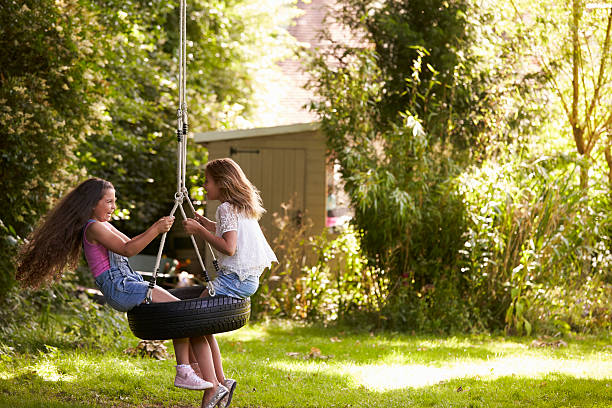 Two Girls Playing Together On Tire Swing In Garden Two Girls Playing Together On Tire Swing In Garden swing stock pictures, royalty-free photos & images