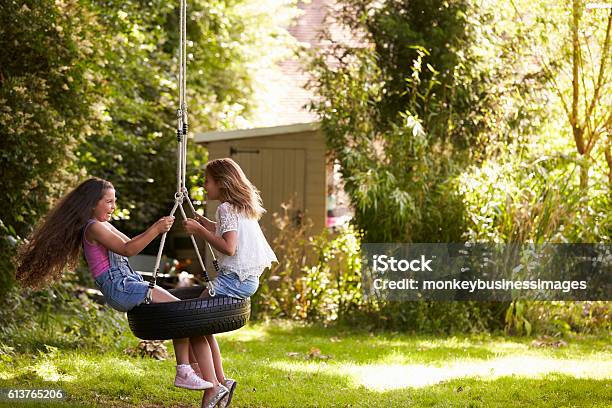 Two Girls Playing Together On Tire Swing In Garden Stock Photo - Download Image Now - Swing - Play Equipment, Child, Swinging