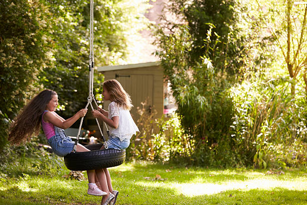 two girls playing together on tire swing in garden - tire swing imagens e fotografias de stock