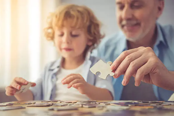 Handsome grandpa and grandson are doing puzzle and smiling while spending time together at home