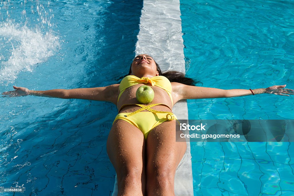 Woman with green apple and measuring tape on wet belly. Young fit woman brunette beauty with yellow bikini rests laying on wet poolside marble with a green apple and measuring tape on her wet belly. Lifestyle and relaxation concept Flat - Physical Description Stock Photo