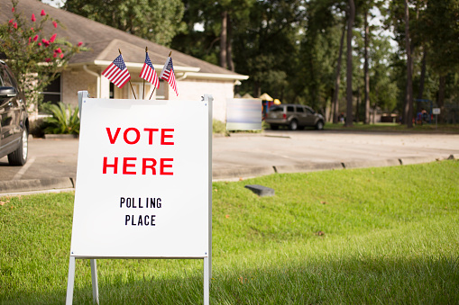 'Vote Here, Polling Place' sign outside of a local, public polling location in USA.  American flags top the sign.  Parking lot and a public community center building in background. The USA elections are held in November each year.  No people.  SIgn created by photographer.