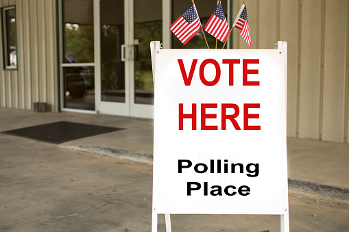 'Vote Here, Polling Place' sign outside of a local, public polling location, which is a public church in USA.  American flags top the sign.  Entrance and public community center building in background. The USA elections are held in November each year.  No people.  Sign created by photographer.