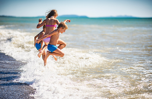 Three kids - a girl and two boys  - are having fun in sea.  Kids are jumping over a wave into the sea. They are caught mid air flying into the sea fun.