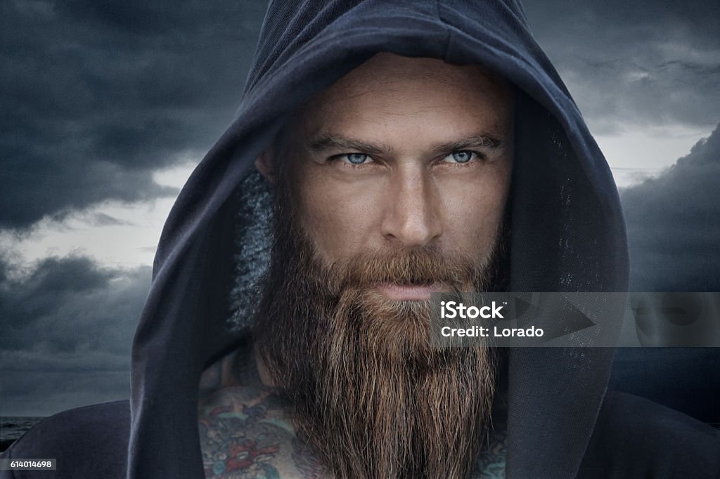 Hooded bearded tattooed male in fantasy cloudy seascape setting - Royaltyfri Män Bildbanksbilder