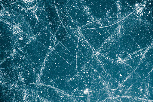 Ice background covered with skid marks going in various directions. The color of the ice changes from place to place. It is a darker shade of blue on the edges and becomes lighter, nearing the shade of white, as it reaches the upper part of the image. The skid marks from skating shoes are white.