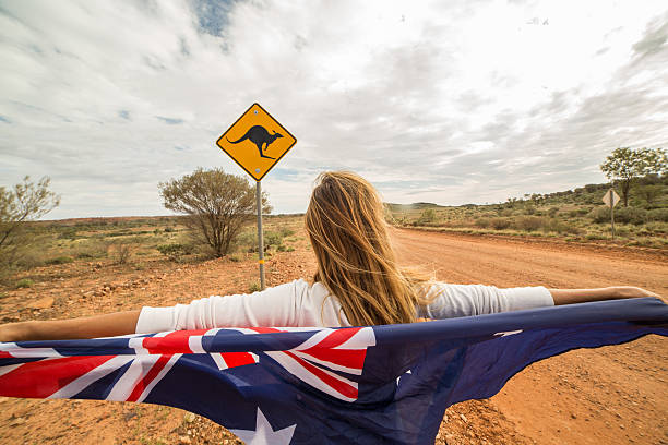 Female holds Australian flag neat Kangaroo sign Cheerful young woman on the road standing next to a kangaroo warning sign, Australia. She is holding an Australian flag in the air. kangaroo crossing sign stock pictures, royalty-free photos & images