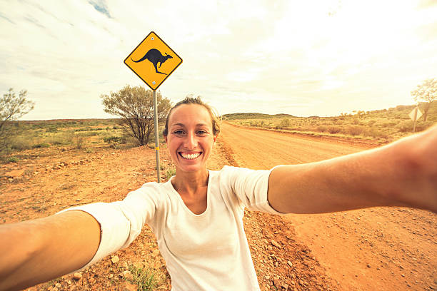 Selfie of young woman in Australia standing near kangaroo sign Cheerful young woman takes a selfie portrait on the road standing next to a kangaroo warning sign, Australia. kangaroo crossing sign stock pictures, royalty-free photos & images