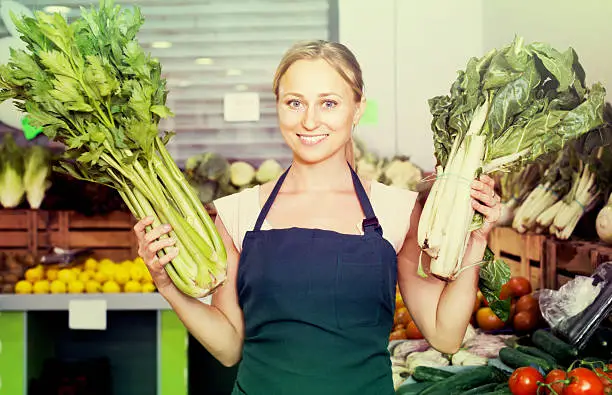 Portrait of young positive woman working showing fresh green celery and lettuce