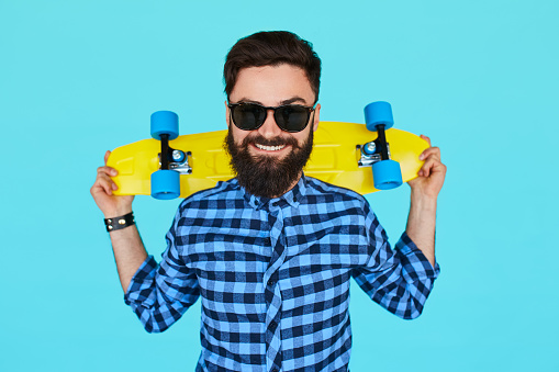 Young hipster man holding a bright vibrant yellow skateboard over colorful blue background. Young male in plaid shirt smiling on color wall