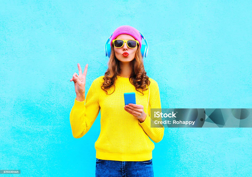 Fashion pretty woman listening music in headphones with smartphone colorful Fashion pretty sweet carefree woman listening music in headphones with smartphone wearing a colorful pink hat yellow sweater sunglasses over blue background Vibrant Color Stock Photo