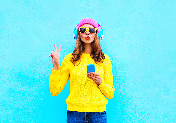 Fashion pretty woman listening music in headphones with smartphone colorful Fashion pretty sweet carefree woman listening music in headphones with smartphone wearing a colorful pink hat yellow sweater sunglasses over blue background hipster fashion stock pictures, royalty-free photos & images
