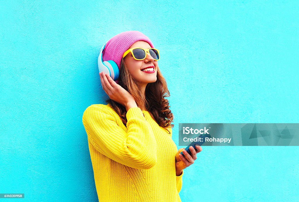 Fashion girl listening to music in headphones with smartphone colorful Fashion pretty sweet carefree girl listening to music in headphones with smartphone wearing a colorful pink hat yellow sunglasses sweater over blue background Music Stock Photo