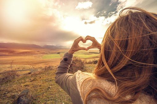 Young woman standing on a hill makes a heart shape finger frame to the spectacular landscape on the background.