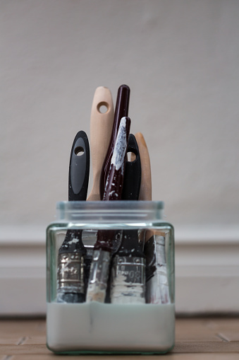 Various paintbrushes soaking in jar of white spirit on a tiled floor. Skirting board visible in background.