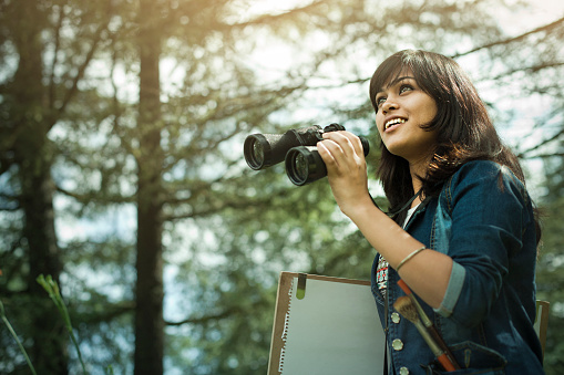 Outdoor image of a late teen female artist holding binocular and walking in nature with drawing board, art materials and tools while searching a subject to paint and looking at view. One person, waist up, horizontal composition with copy space.