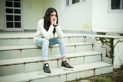 Outdoor image of Asian, sad girl looking down, thinking and sitting on steps by holding her head. She is in jeans and white sweater. One person, full length, horizontal composition with copy space.