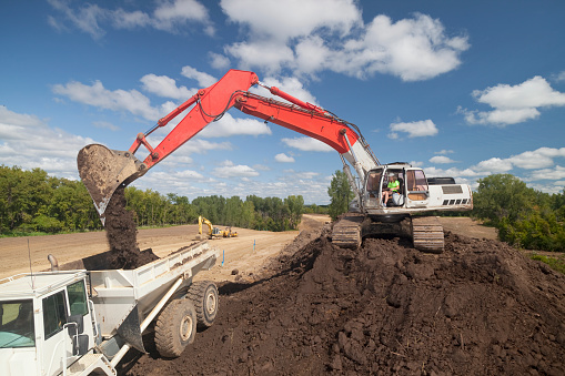 A female or woman construction worker operating an excavator on top a mound of dirt, unloading the dirt into a dump truck.
