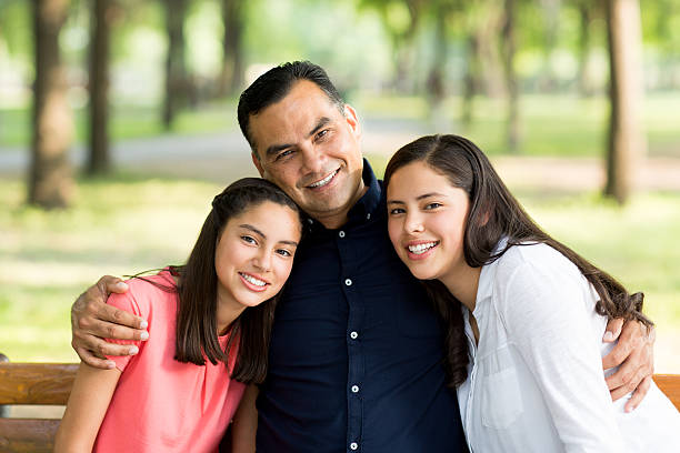 Latin father embracing his daughters and smiling at camera A mature latin father embracing his two teen daughters, sitting on a bench and smilng at the camera in a horizontal waist up shot outdoors. single father stock pictures, royalty-free photos & images