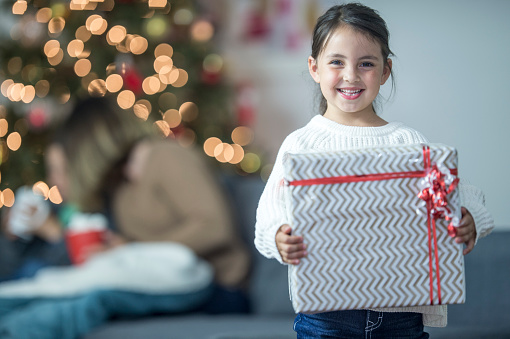 A little girl is happily standing with her Christmas present and is smiling and looking at the camera.