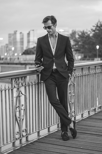 Young handsome hipster businessman standing on the bridge, and texting on mobile phone. Black and white photo.