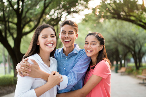 Fun latin teenage brother and sisters embracing, looking at the camera and smiling in a horizontal waist up shot outdoors.