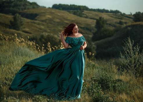 Beautiful young woman in a green dress, walking among steep hills. Long skirt flutters in the wind. Fabulous, mysterious forest. The picturesque nature. Fashion tinted. Creative color.