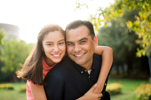 A latin teen girl embracing her father, looking at the camera and smiling in a horizontal waist up shot outdoors.