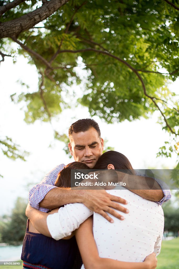 Latin father praying with teen daughters under tree A latin father praying with his teen daughters under a tree in a vertical waist up shot outdoors. Embracing Stock Photo