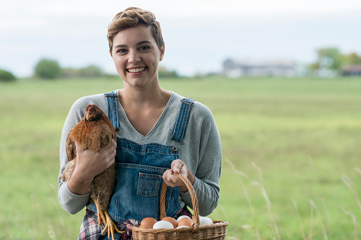 A farmer looks at one of her chickens while holding it and some eggs in a basket. She stands with a field in the background.
