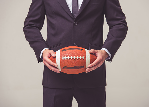Cropped image of handsome young businessman in suit holding a football ball, on a gray background
