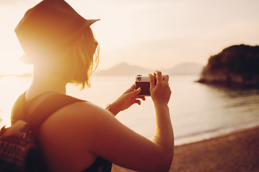 Woman traveler with camera taking photo near sea at sunset