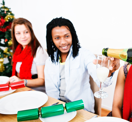 A group of young friends sharing Christmas dinner;  one pours champagne as a smiling guest holds up his glass to receive it..