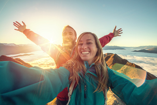 Young couple hiking stand on a mountain peak above the clouds and take a selfie portrait, sun shining over the mountains. Beautiful sunbeam effect making an idyllic landscape.