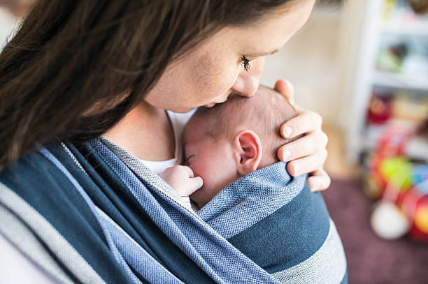 Close up of unrecognizable young mother with her newborn baby son in sling at home, kissing him
