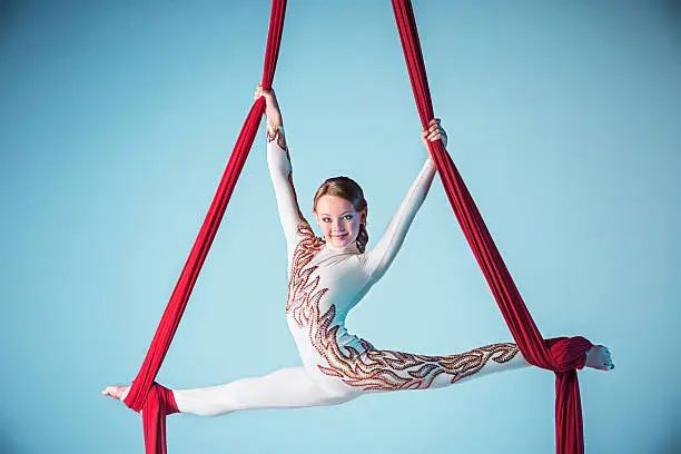 Graceful gymnast performing aerial exercise with red fabrics on blue background