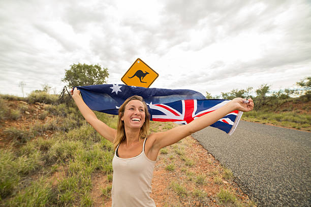 Female holds Australian flag neat Kangaroo sign Cheerful young woman on the road standing next to a kangaroo warning sign, Australia. She is holding an Australian flag in the air. kangaroo crossing sign stock pictures, royalty-free photos & images