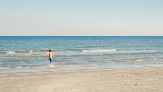 Rear view of a boy running into the waves in the ocean. He is wearing a swimsuit. Copy space is available.